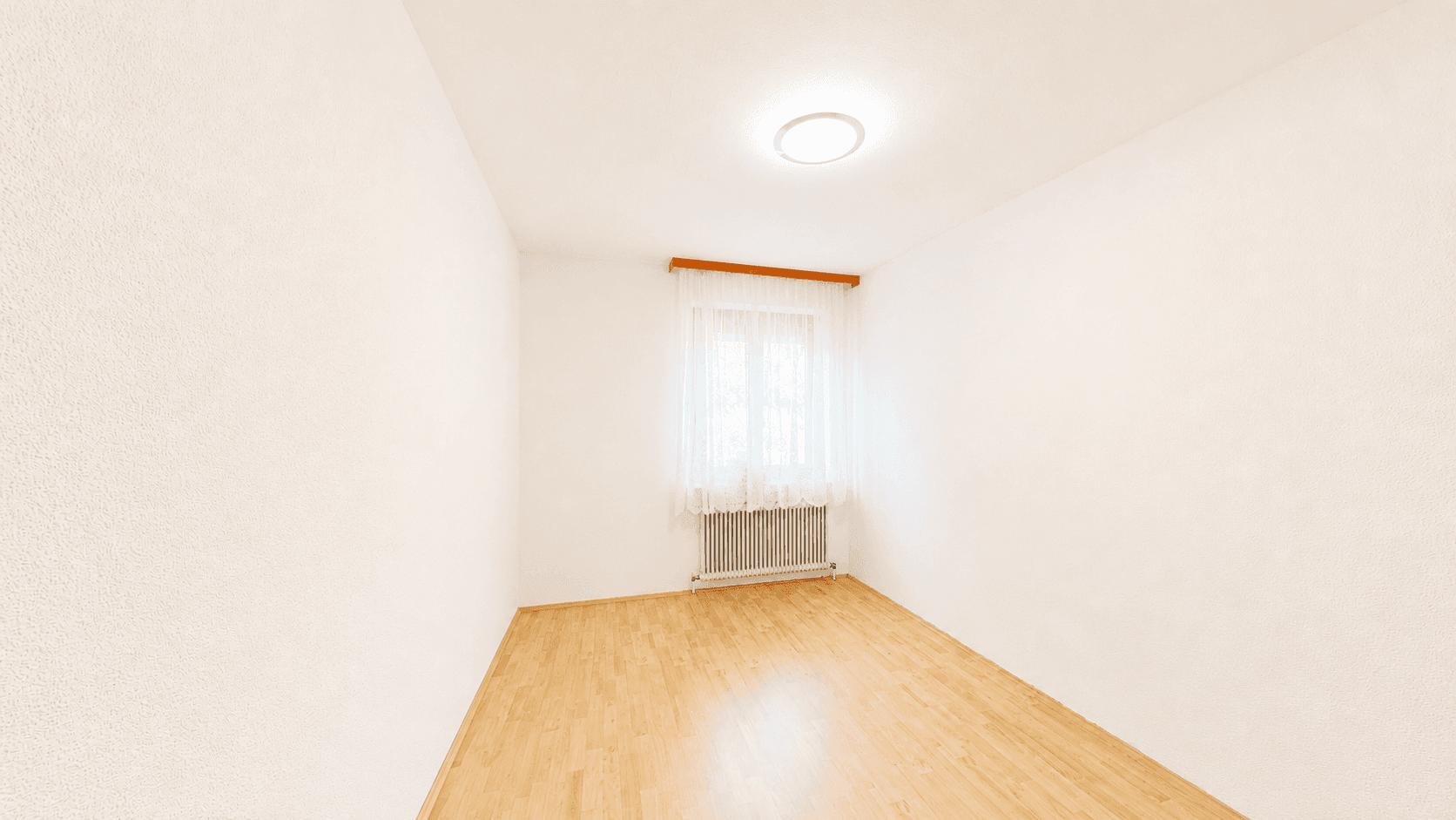 Schlichter, leerer Raum mit Laminatboden, weißen Wänden und einem Fenster mit hellen Gardinen.