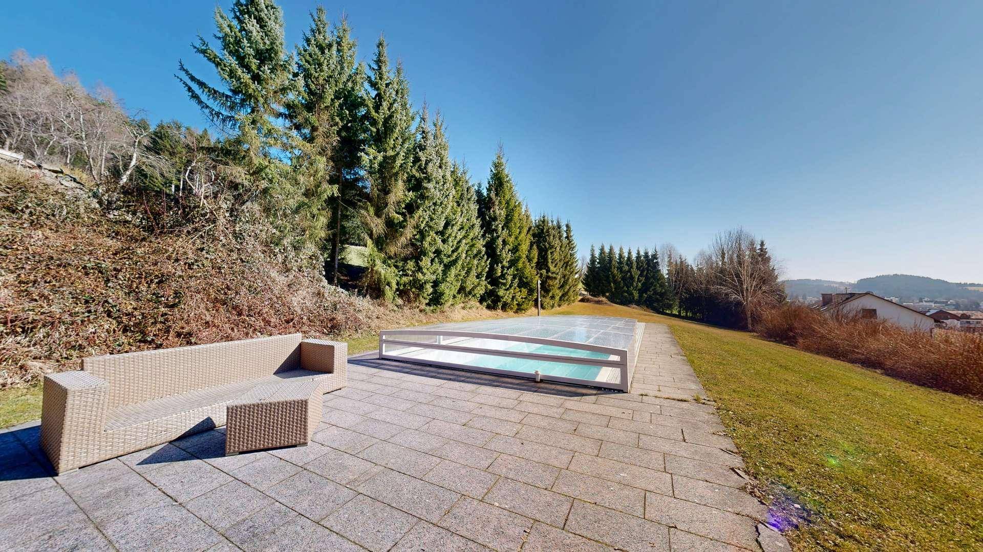 Außenbereich mit überdachtem Swimmingpool, gepflasterter Liegefläche und Sitzgelegenheit im Grünen.