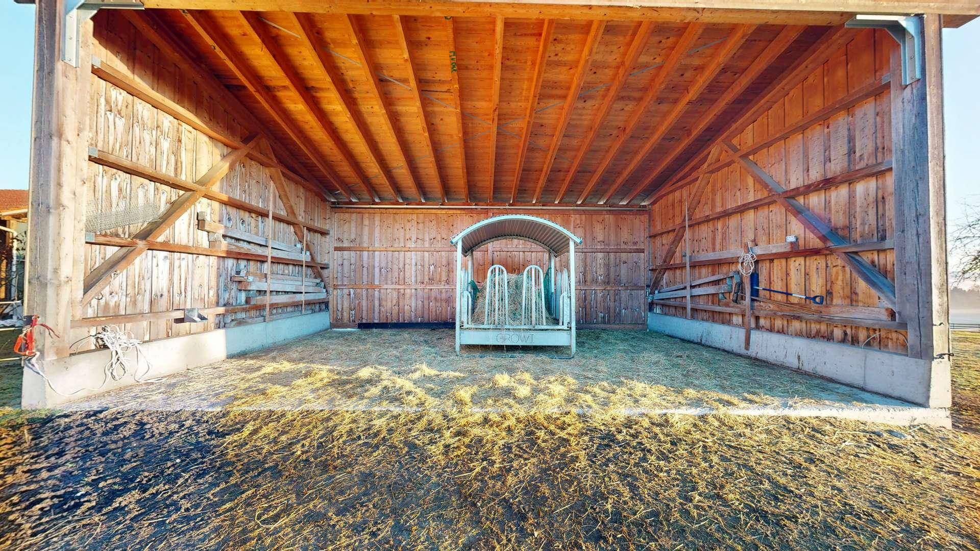 Geräumiger Holzunterstand mit Heuraufe und sichtbaren Dachbalken.