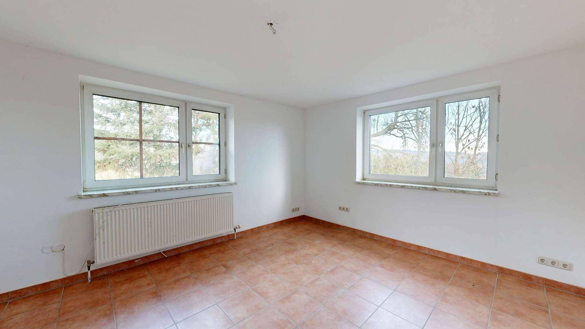 Heller Raum mit zwei Fenstern, Fliesenboden und Heizkörper unter den Fenstern.