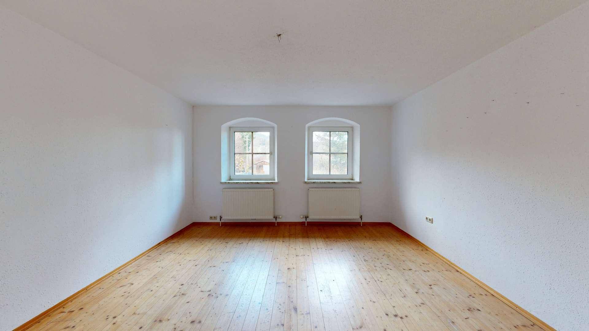 Leerer Raum mit Holzdielenboden und zwei Fenstern mit Blick nach draußen.