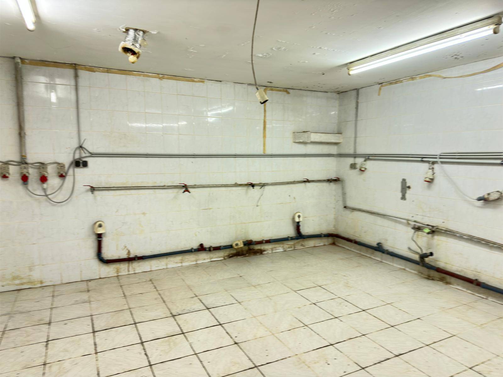 Werkstattraum mit weißen Fliesenwänden, vielen sichtbaren Rohren und einfacher Neonbeleuchtung an der Decke.