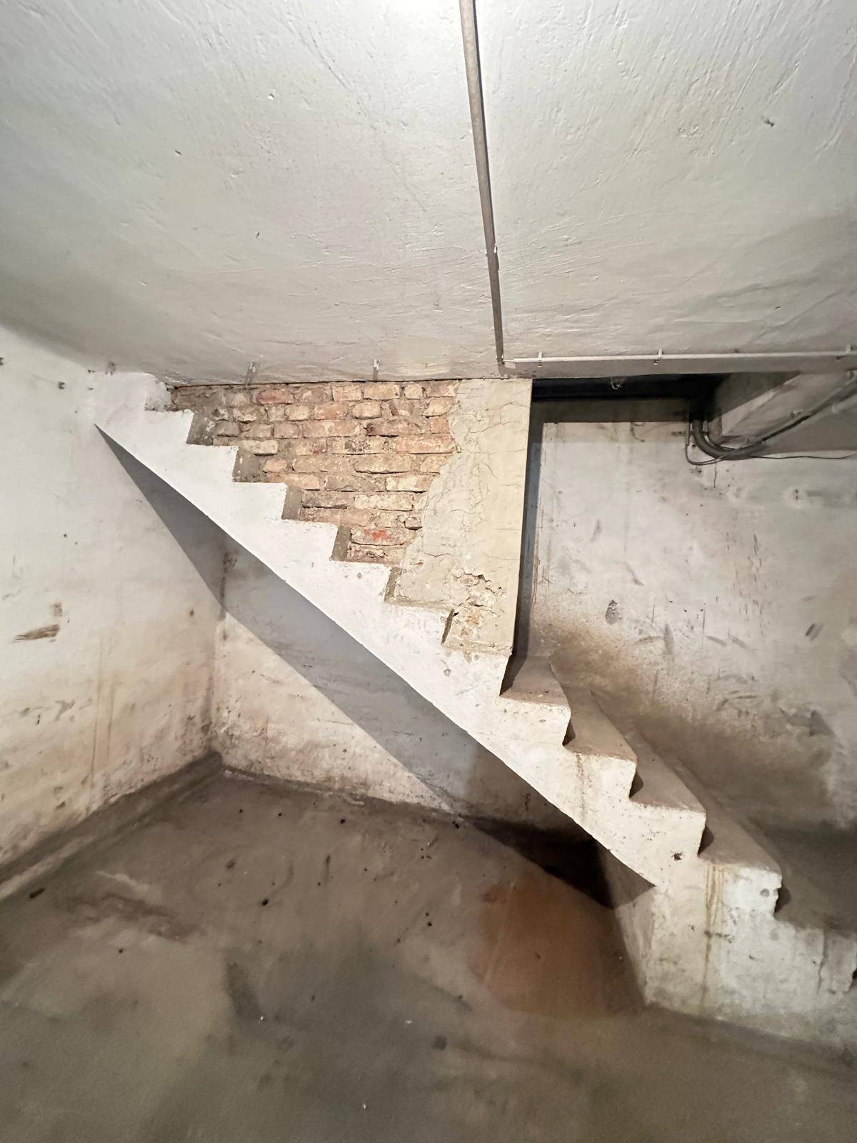 Betonierte Treppe führt in einen Kellerraum mit rauen Wänden, sichtbaren Ziegeln und schlichter Decke.