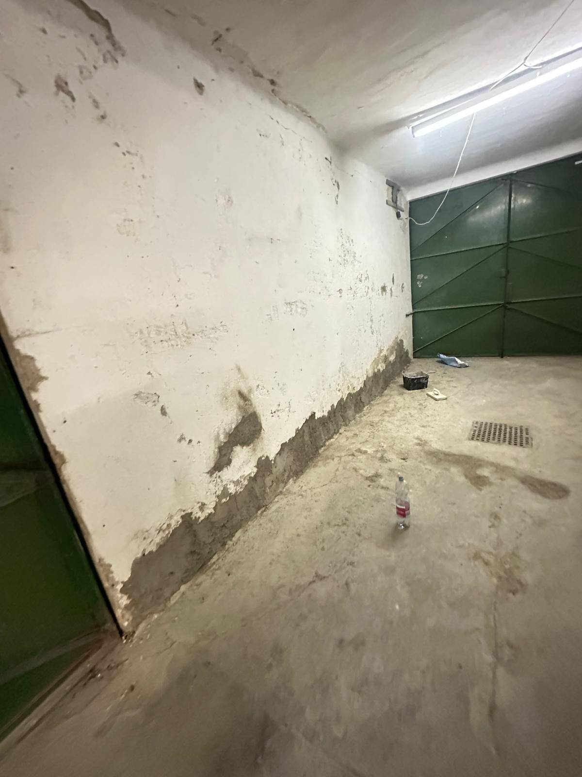 Innenraum einer Garage mit Betonboden, grünem Tor und sichtbaren Abnutzungsspuren an den Wänden und Decken.