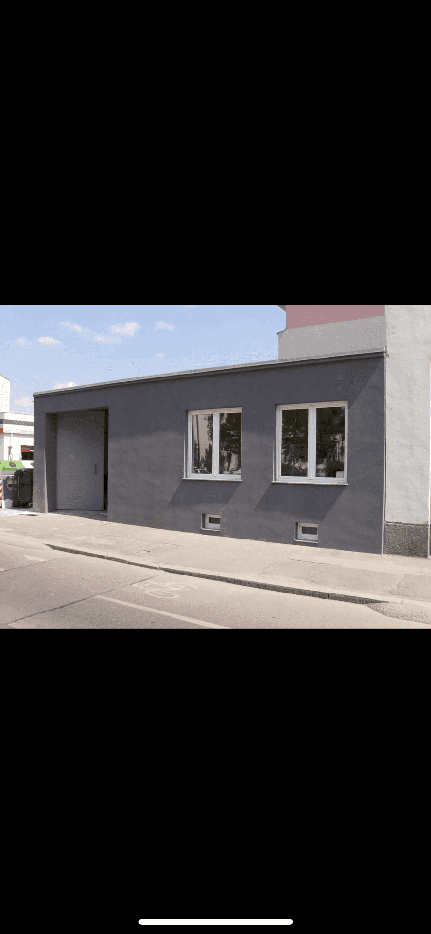 Graue Fassade eines funktionalen Gebäudes mit zwei Fenstern und einem großen Tor, direkt an einer Straße gelegen.