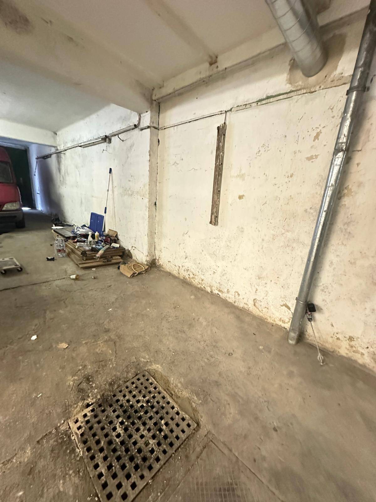 Innenansicht einer Garage mit Betonboden, Abflussgitter und sichtbaren Rohren an den Wänden und Decken.