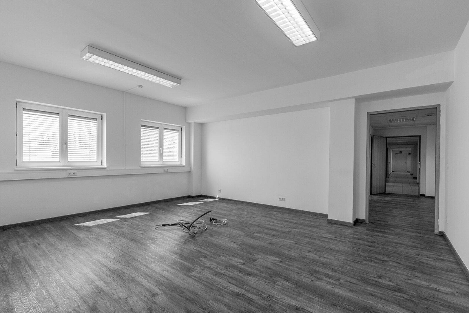 Leeres Büro mit Laminatboden, weißen Wänden und natürlichem Lichteinfall durch die Fenster.