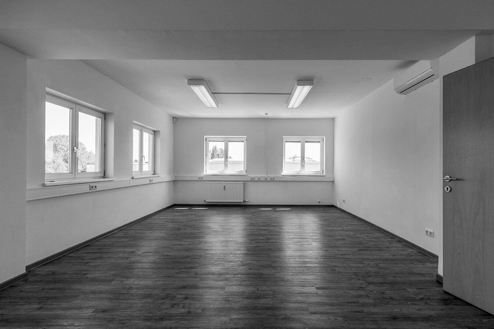 Geräumiges Büro mit mehreren Fenstern, Laminatboden und einer Klimaanlage an der Wand.
