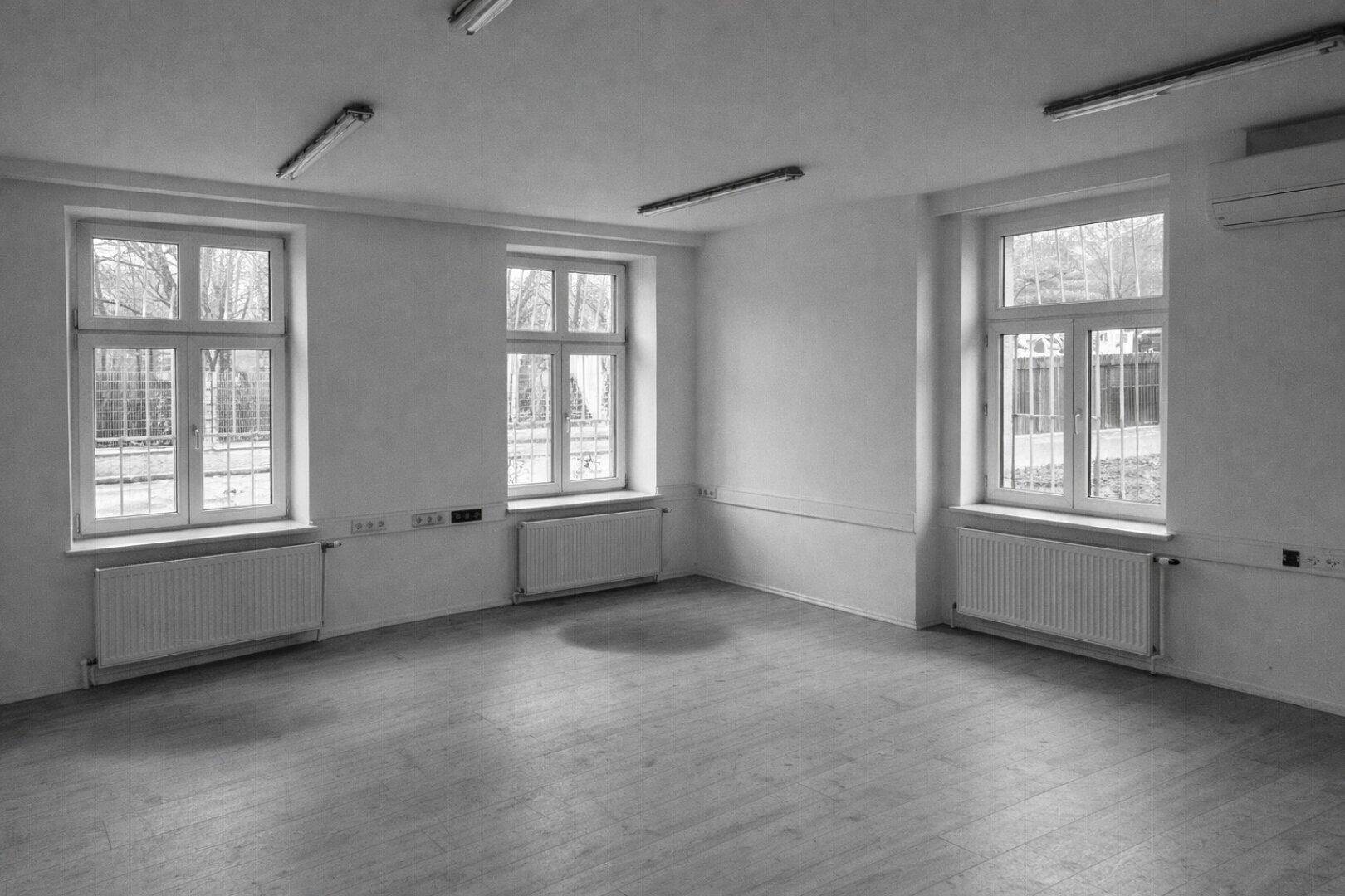 Heller, leerer Raum mit drei großen Fenstern, Heizkörpern und Holzboden.
