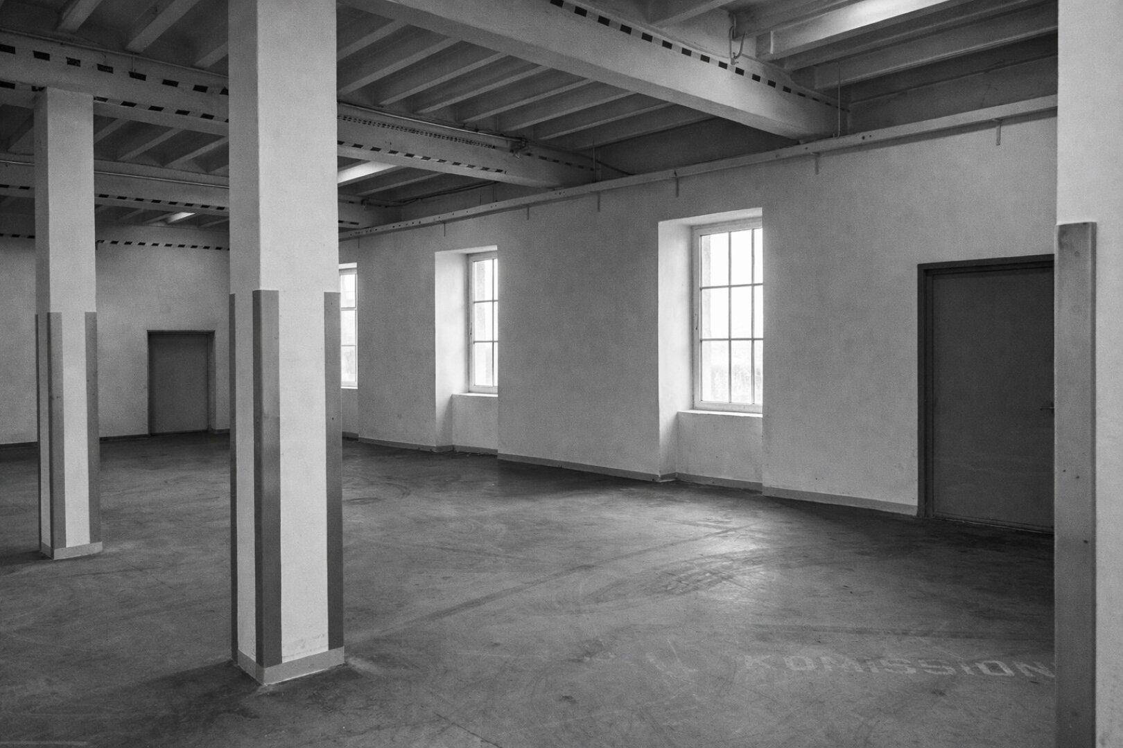Geräumige Halle mit Betonboden, tragenden Säulen und mehreren Fenstern, die viel Licht hereinlassen.