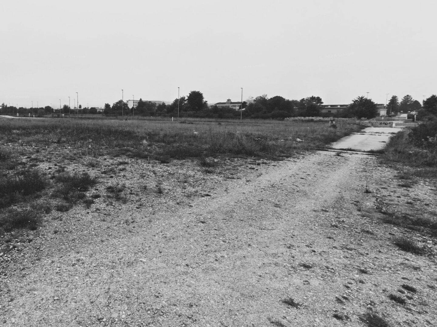 Weitläufiges, brachliegendes Feld mit einem Schotterweg, umgeben von spärlicher Vegetation und entfernten Gebäuden.