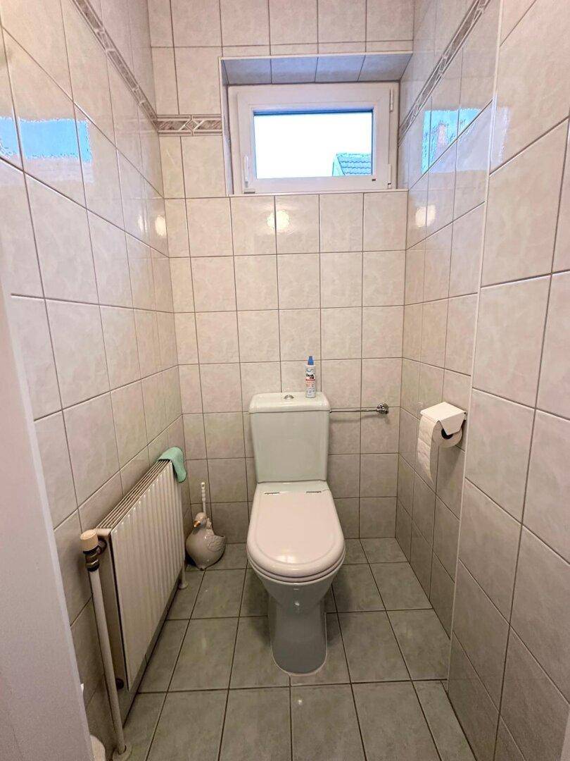 Separate Toilette mit weißen Fliesen, WC und einem kleinen Fenster.