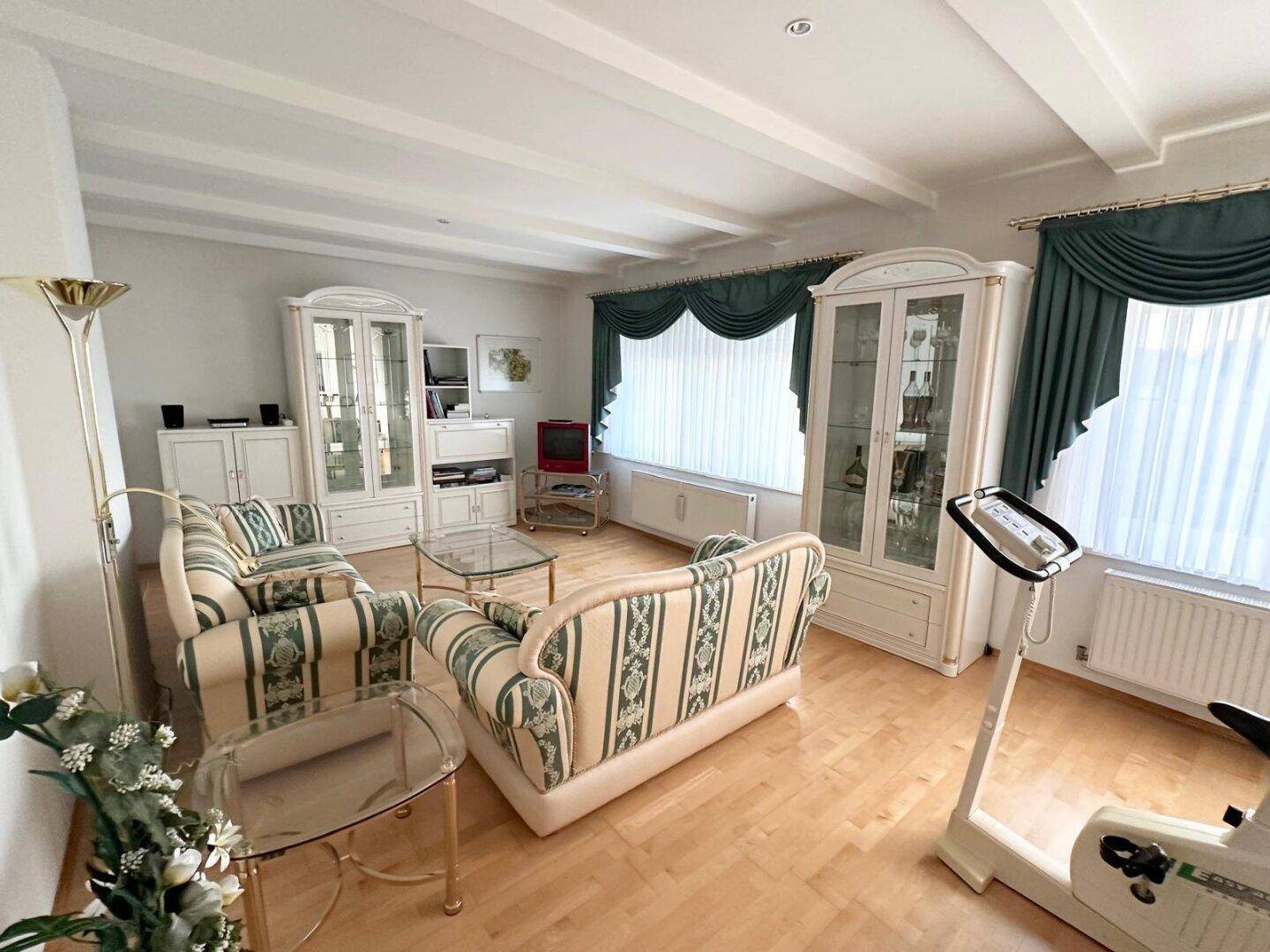 Helles Wohnzimmer mit gemusterten Sofas, weißen Schränken und sichtbaren Holzdeckenbalken.