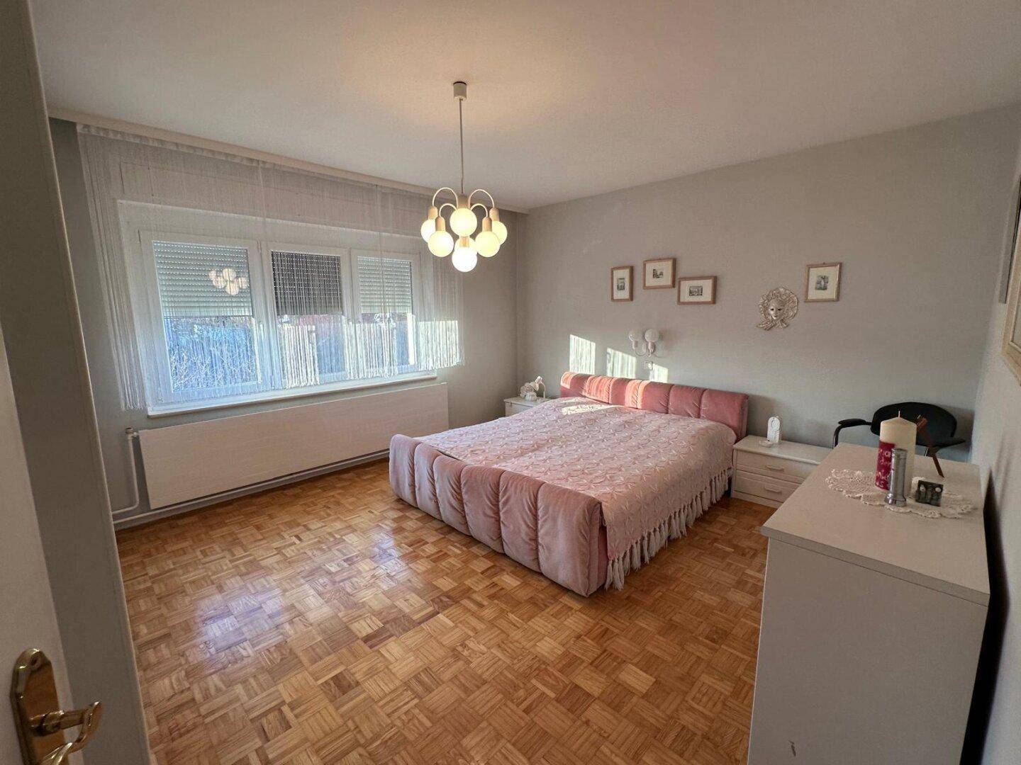 Schlafzimmer mit einem auffälligen rosa Bett, weißen Möbeln und Parkettboden.