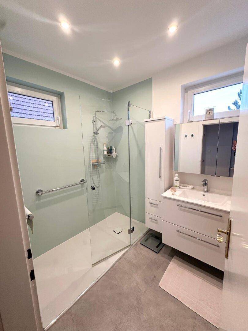 Modernes Badezimmer mit ebenerdiger Dusche, Waschtisch und zwei Fenstern.