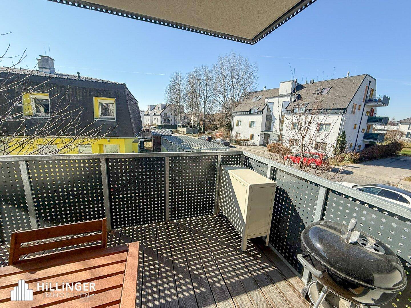 Sonniger Balkon mit Holzboden, Grill und Sitzgelegenheiten, umgeben von Wohngebäuden.
