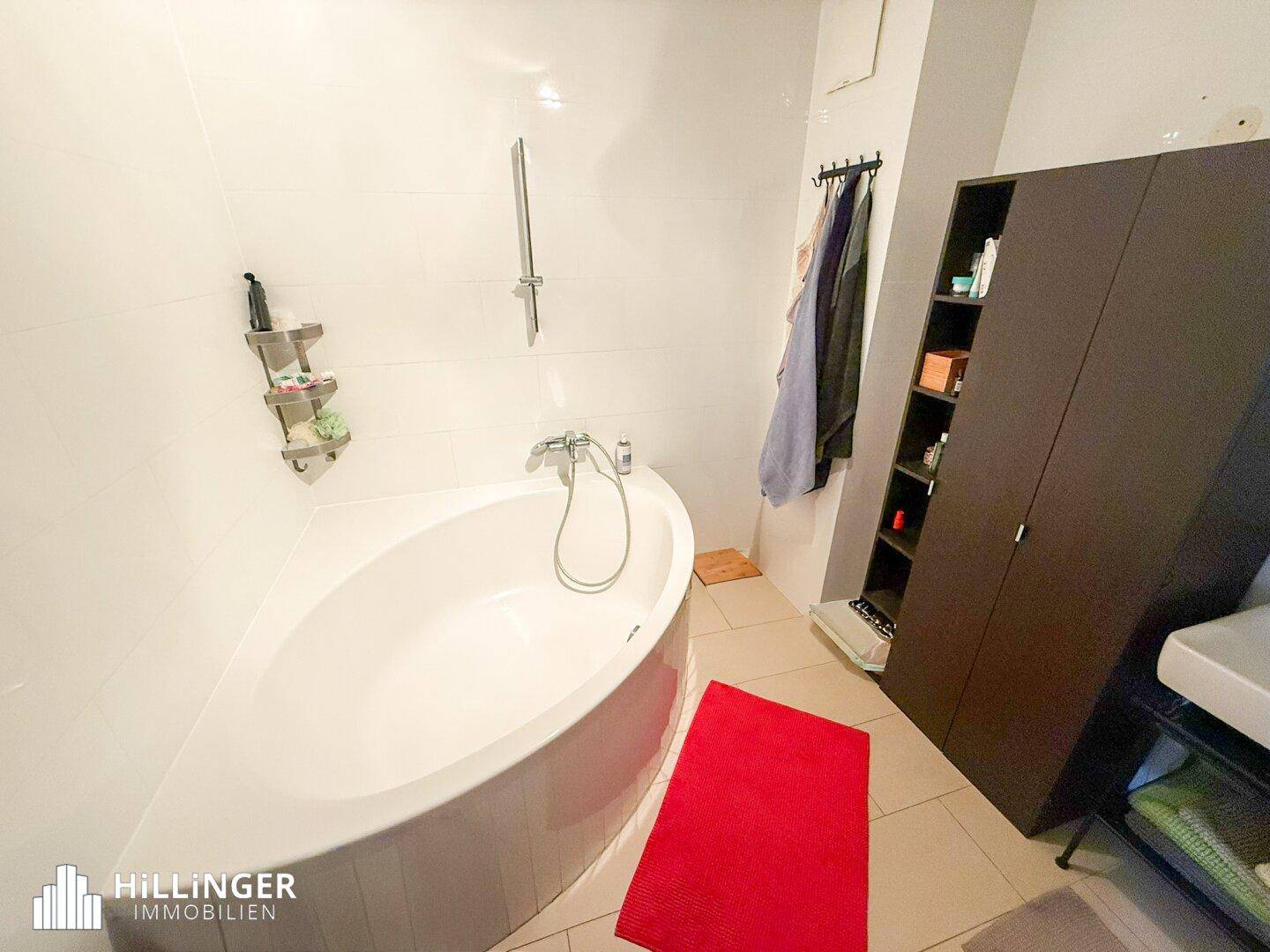 Badezimmer mit weiß gefliesten Wänden, einer Eckbadewanne mit Duschfunktion und Stauraum.