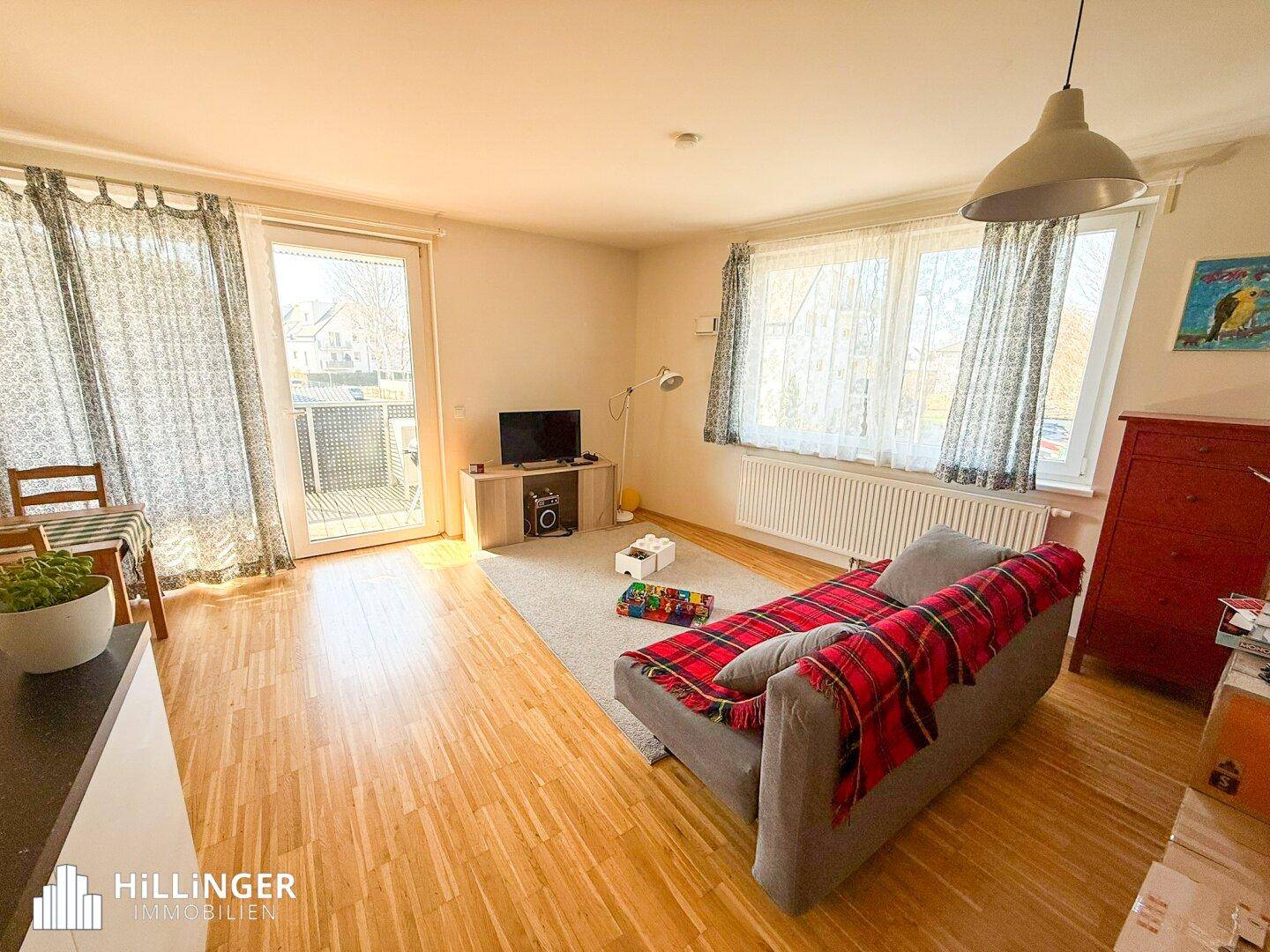 Helles Wohnzimmer mit Holzboden, großem Fenster und Zugang zu einem Außenbereich.