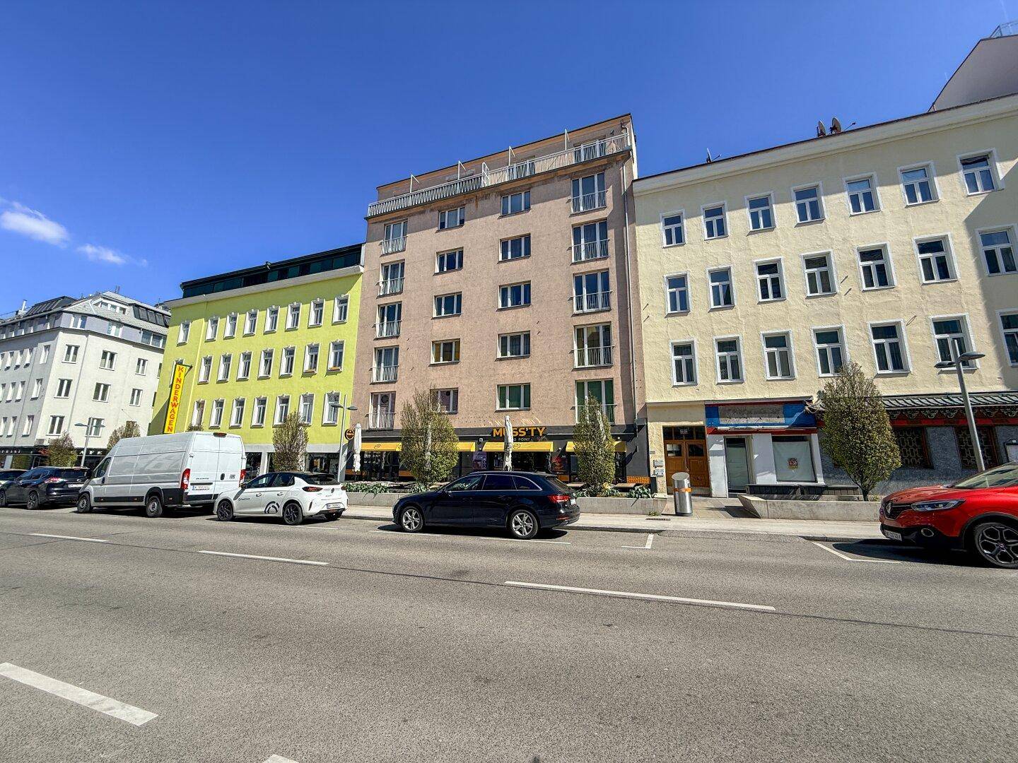 Mehrere Gebäude entlang einer belebten Straße mit parkenden Autos und Geschäften im Erdgeschoss.