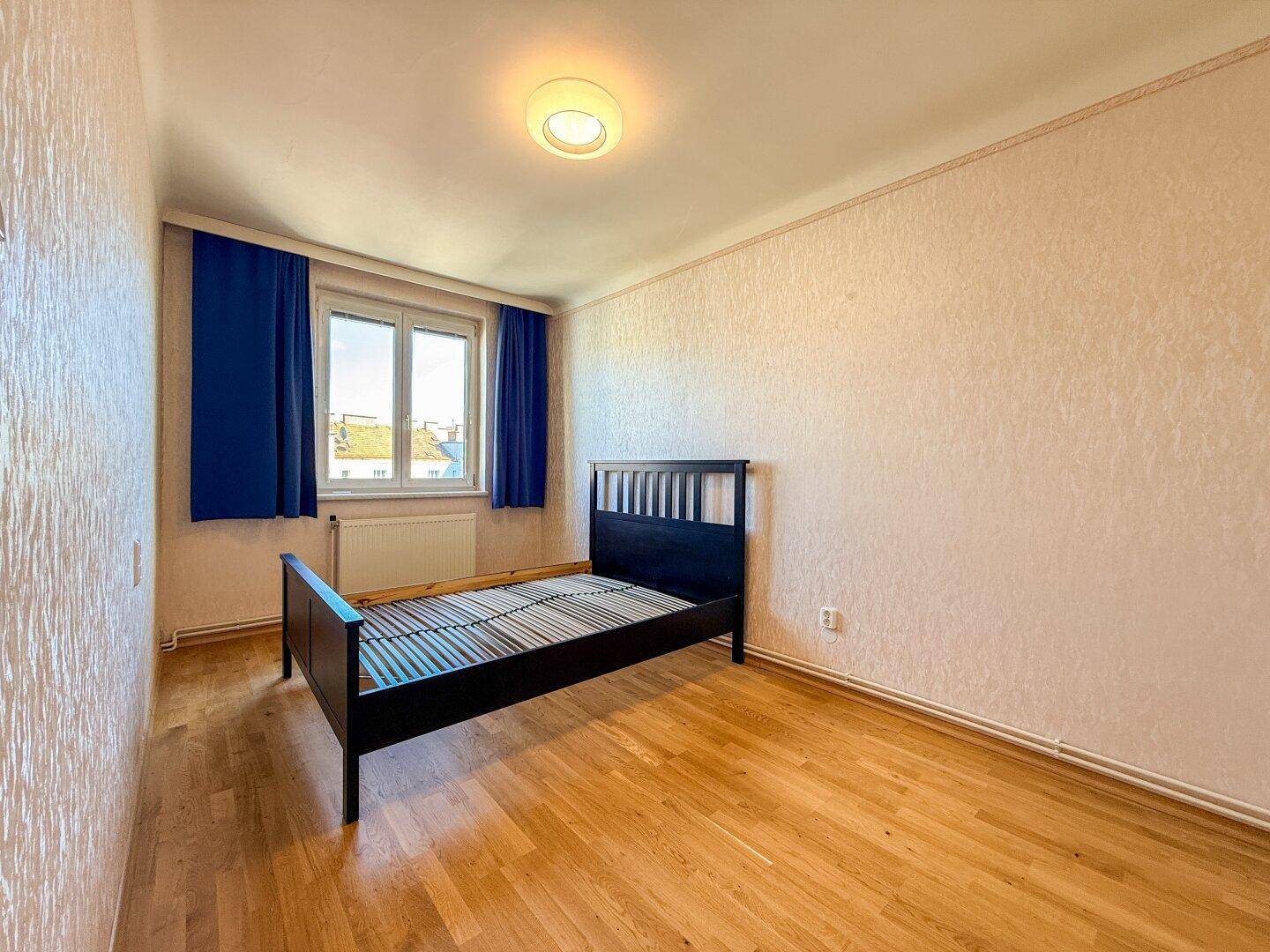 Schlafzimmer mit Parkettboden, blauem Vorhang und einem Fenster, das viel Licht hereinlässt.