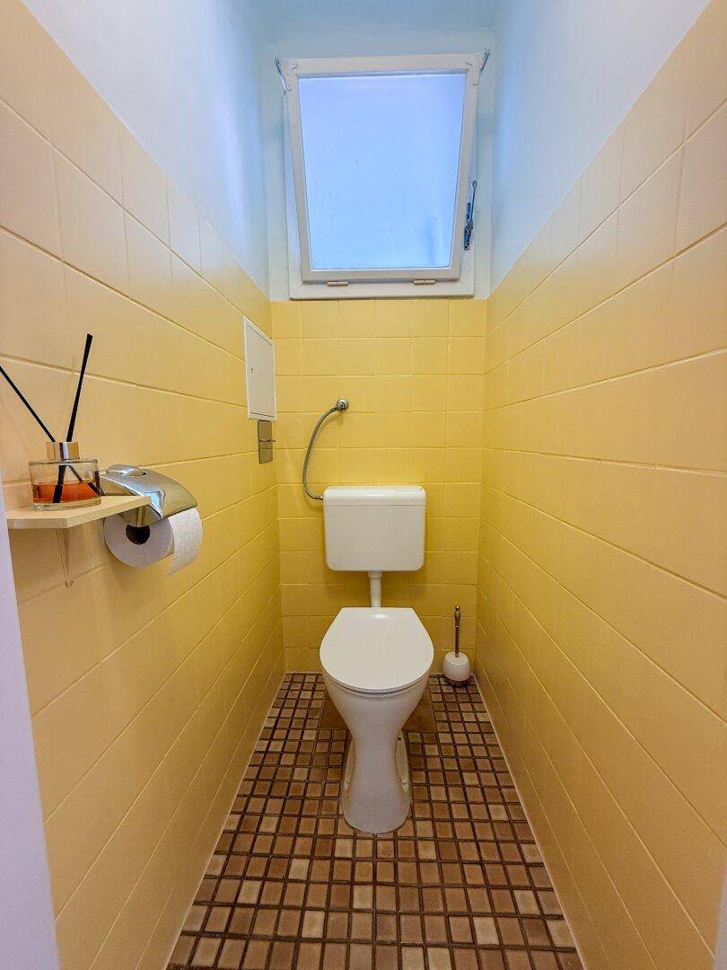 Kleines WC mit gelben Fliesen an den Wänden und einem Fenster für natürliche Belüftung.