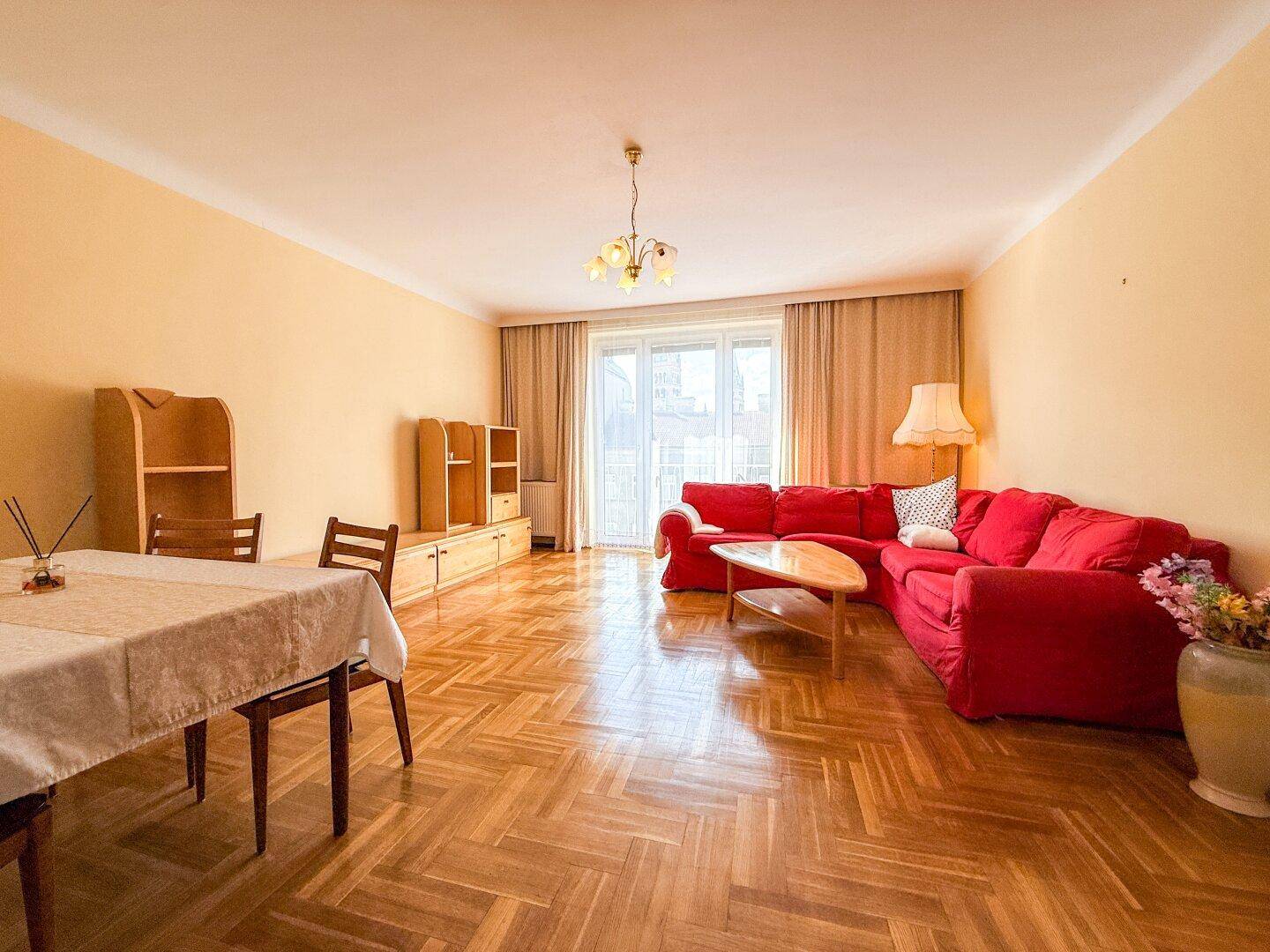 Geräumiger Wohn- und Essbereich mit Parkettboden, rotem Sofa und Zugang zum Balkon.