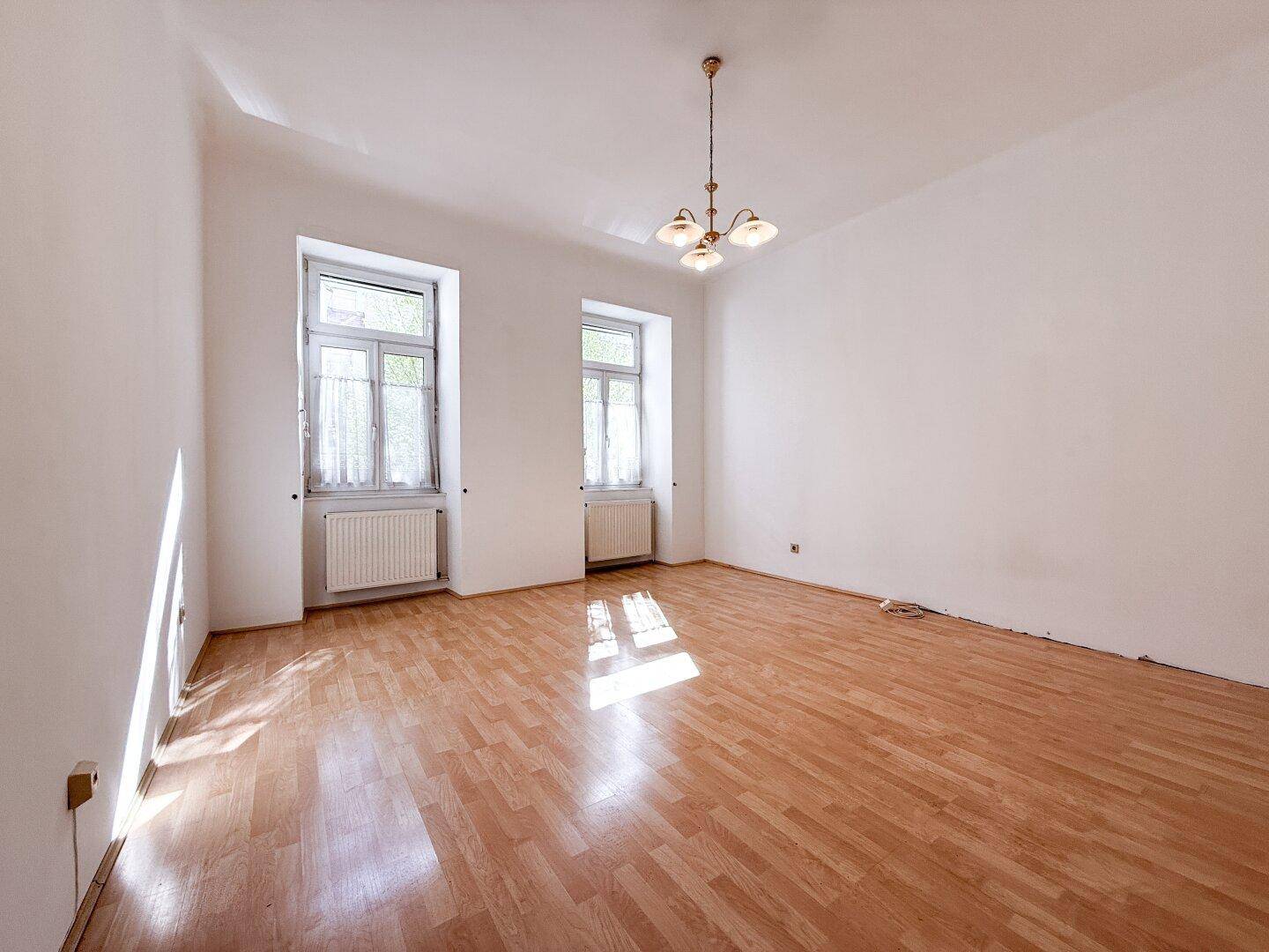 Helles, leeres Wohnzimmer mit Parkettboden und zwei Fenstern, durch die Sonnenlicht fällt.