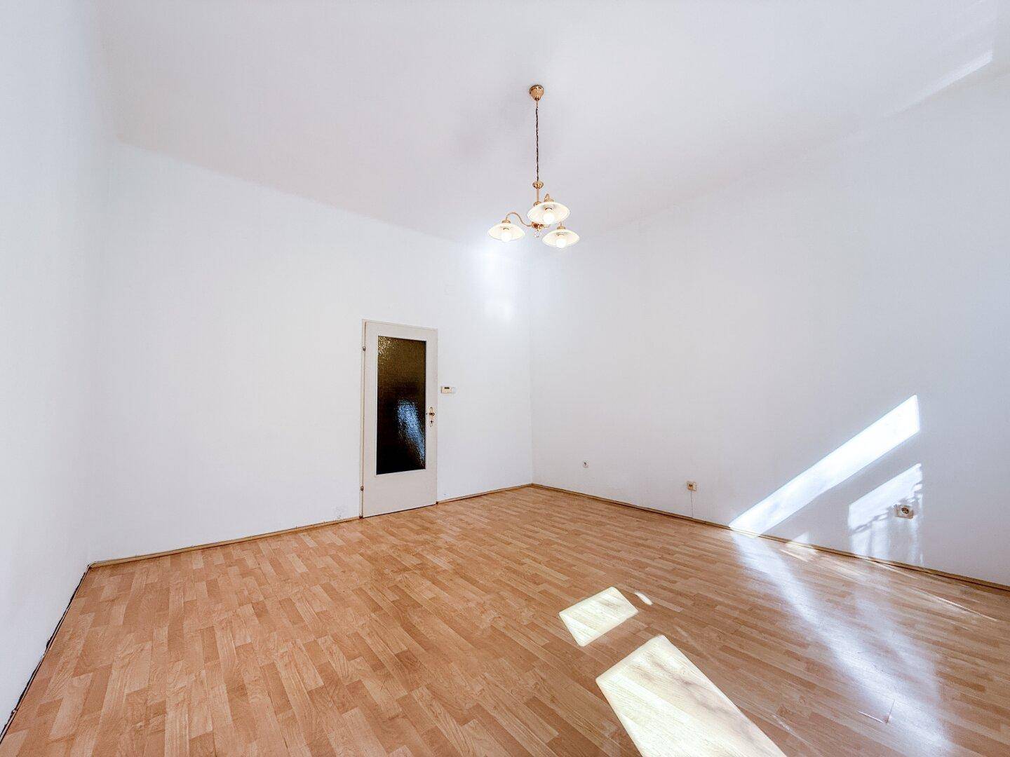 Geräumiges, leeres Wohnzimmer mit Parkettboden und einer Tür, durch die Sonnenlicht fällt.