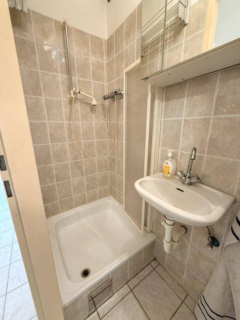 Kleines Badezimmer mit Duschkabine, Waschbecken und hellen Fliesen an Wänden und Boden.