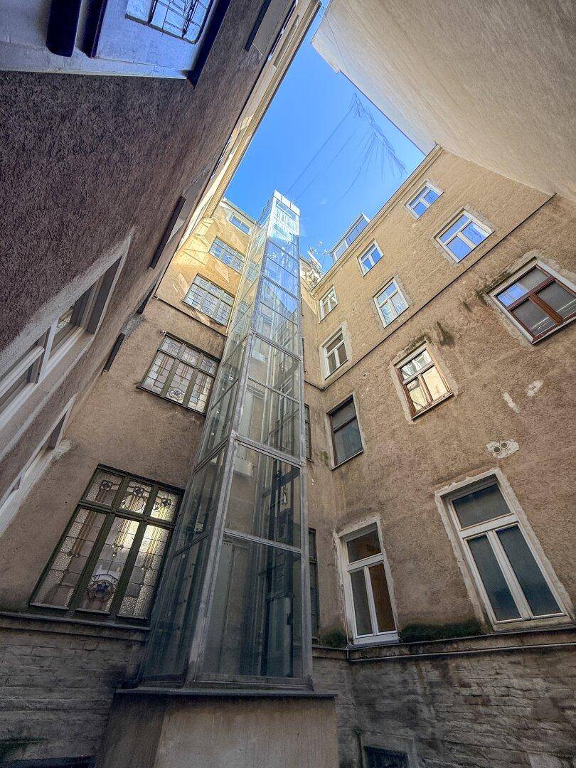 Blick in einen engen Innenhof mit einem modernen Glasaufzug und alten Gebäudefassaden.