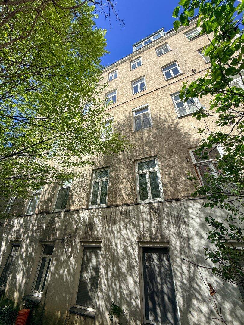 Fassade eines alten Gebäudes, teilweise von grünen Bäumen verdeckt, unter blauem Himmel.