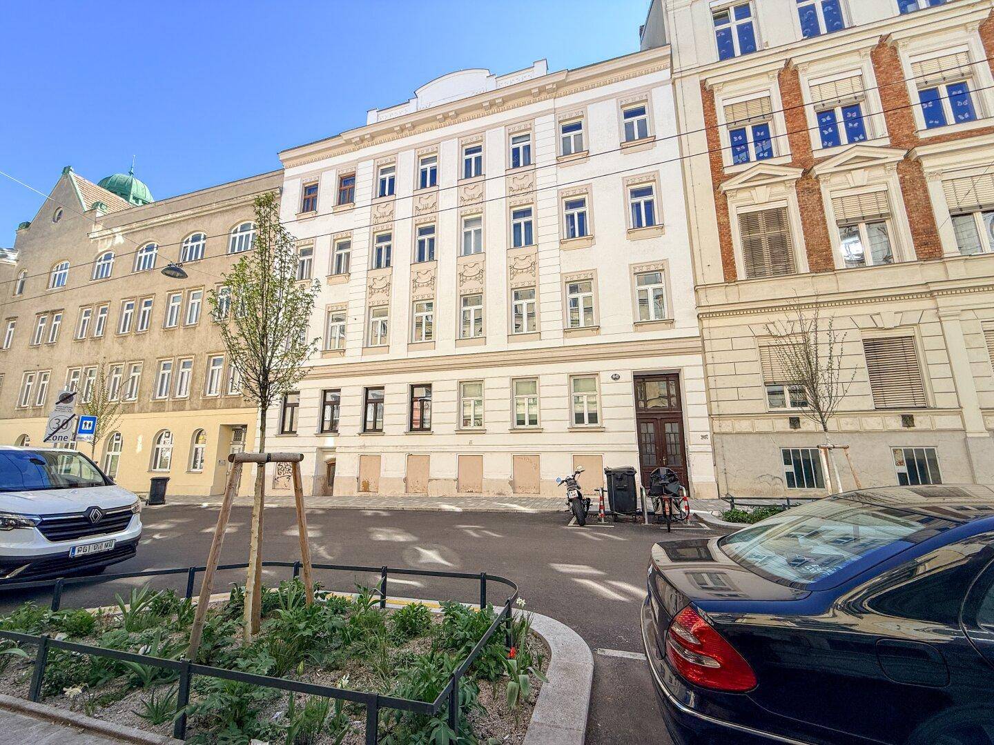 Straßenansicht mit der Gebäudefassade, geparkten Autos und umliegenden Häusern.