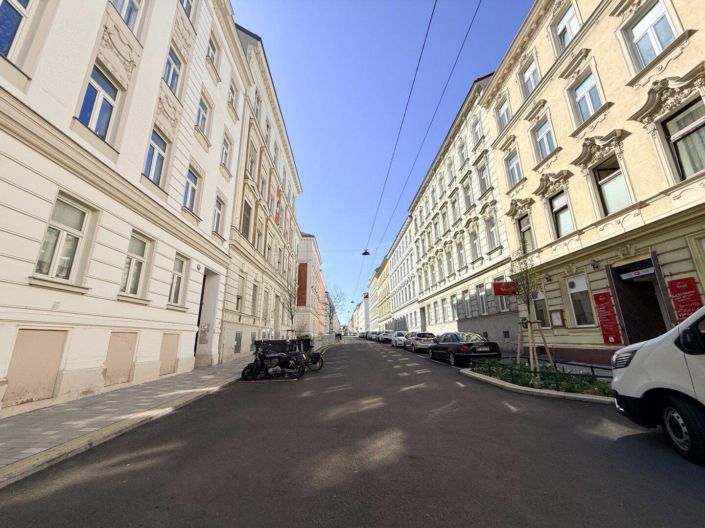 Lange Straßenansicht mit Gebäuden auf beiden Seiten und geparkten Fahrzeugen.