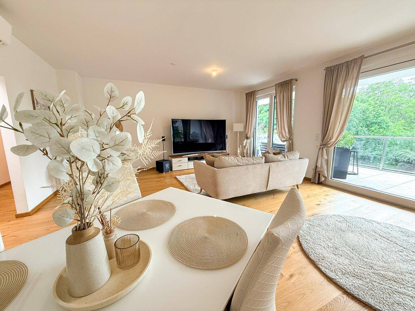 Heller Wohn- und Essbereich mit Holzboden, modernen Möbeln und Zugang zu einem Balkon mit Grünblick.