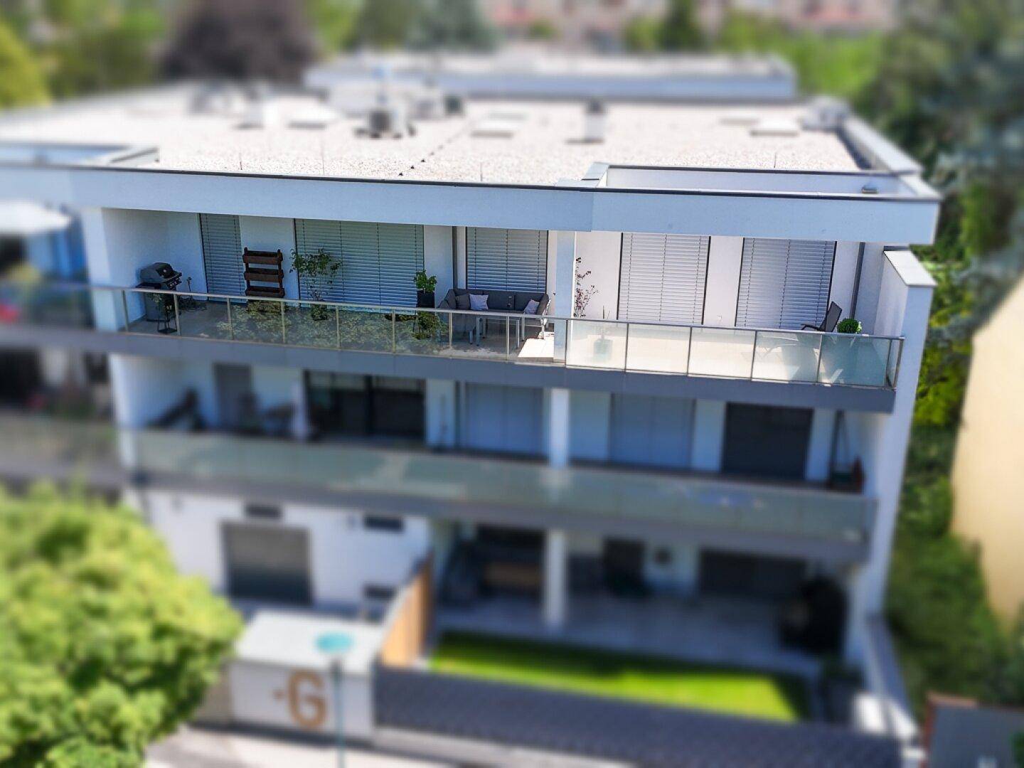 Modernes Mehrfamilienhaus mit mehreren Balkonen und Flachdach, umgeben von Grünflächen und Bäumen.