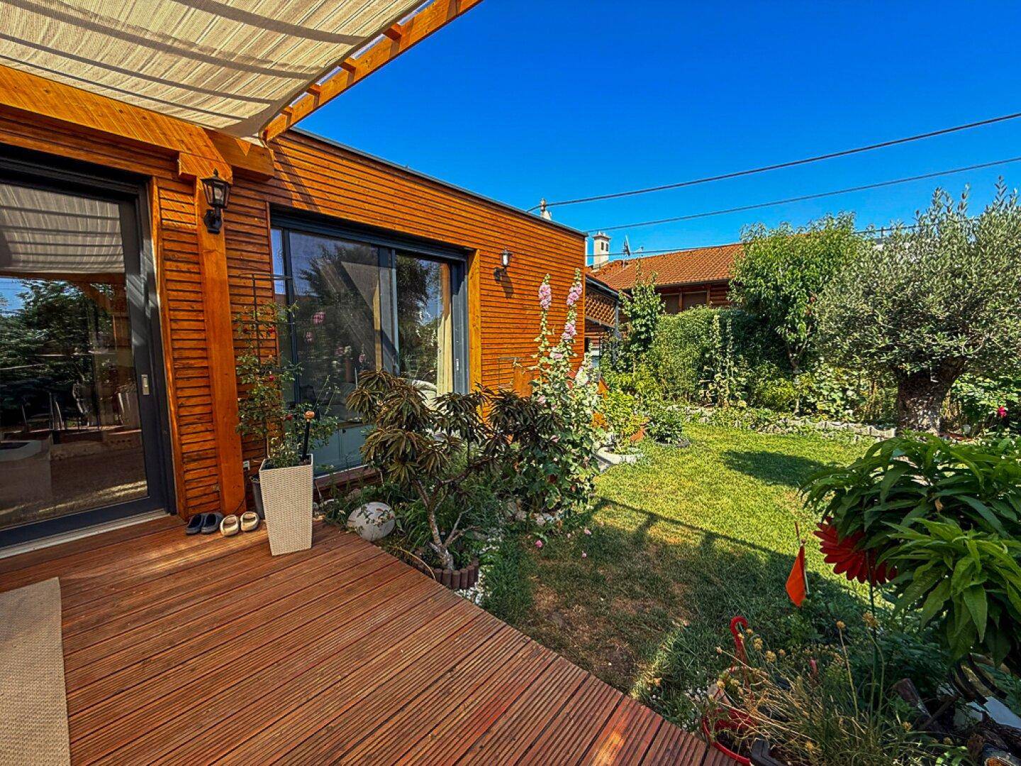 Holzfassade des Hauses mit angrenzender Holzterrasse und einem blühenden, grünen Garten.