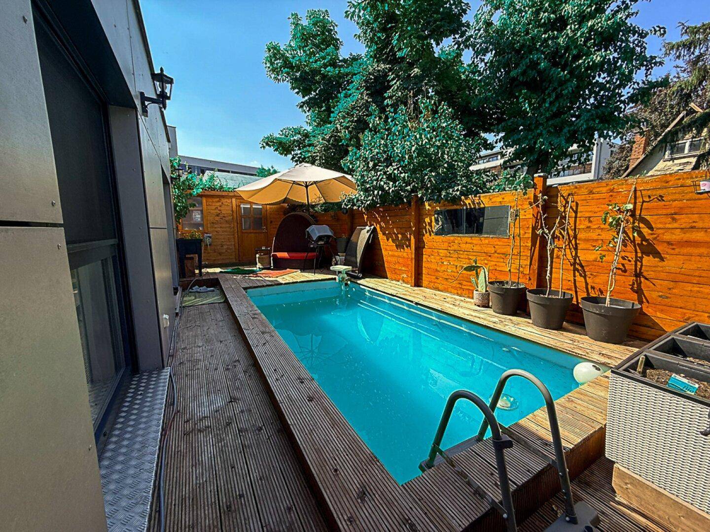 Außenpool mit Holzdeck und Sonnenschirm, umgeben von einem Holzzaun und grüner Bepflanzung.
