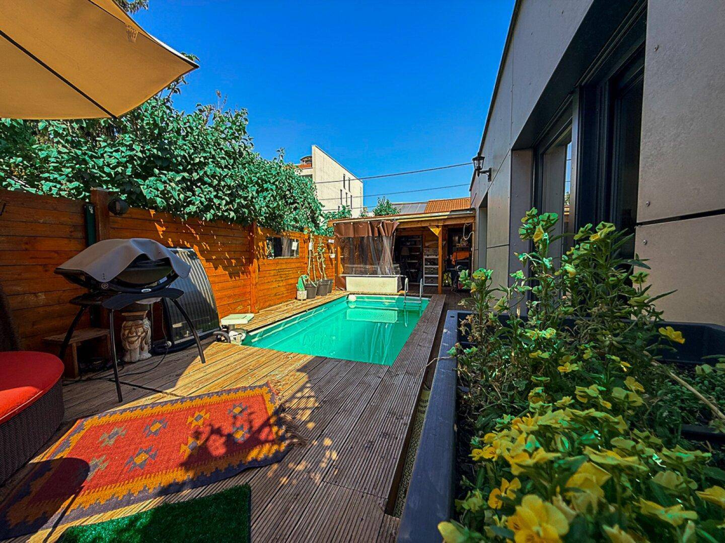 Sonniger Poolbereich mit Holzdeck, umgeben von grünen Pflanzen und einem Holzzaun.