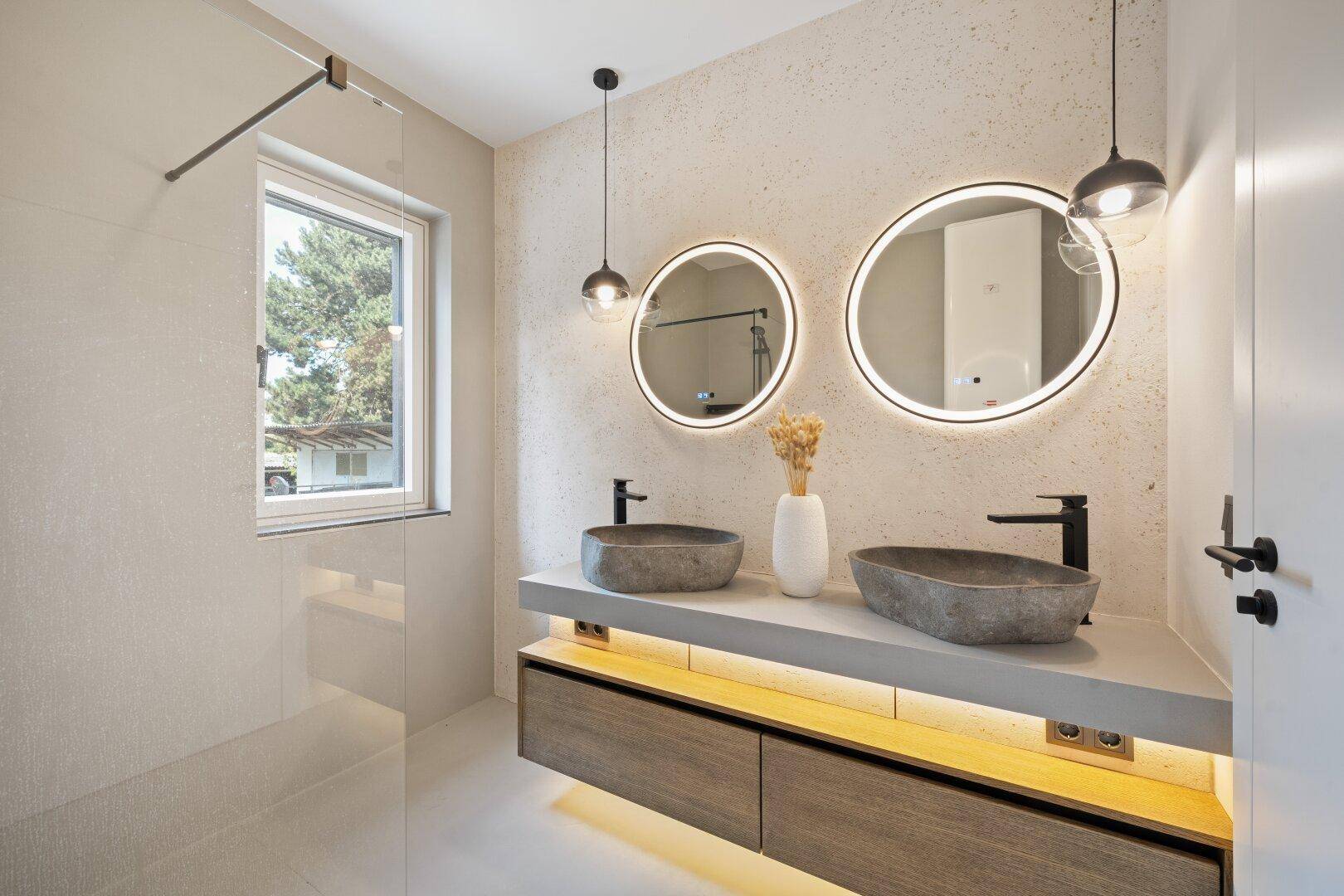 Modernes Badezimmer mit Doppelwaschbecken, beleuchteten Spiegeln und Fenster für Tageslicht.