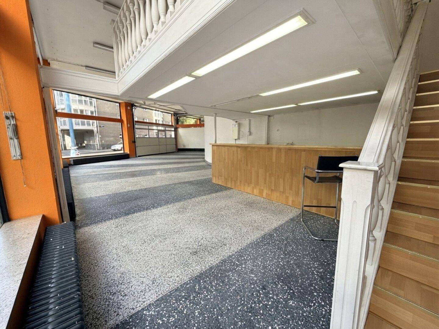 Blick von der Treppe auf den geräumigen Verkaufsraum mit Theke und gestreiftem Bodenbelag.