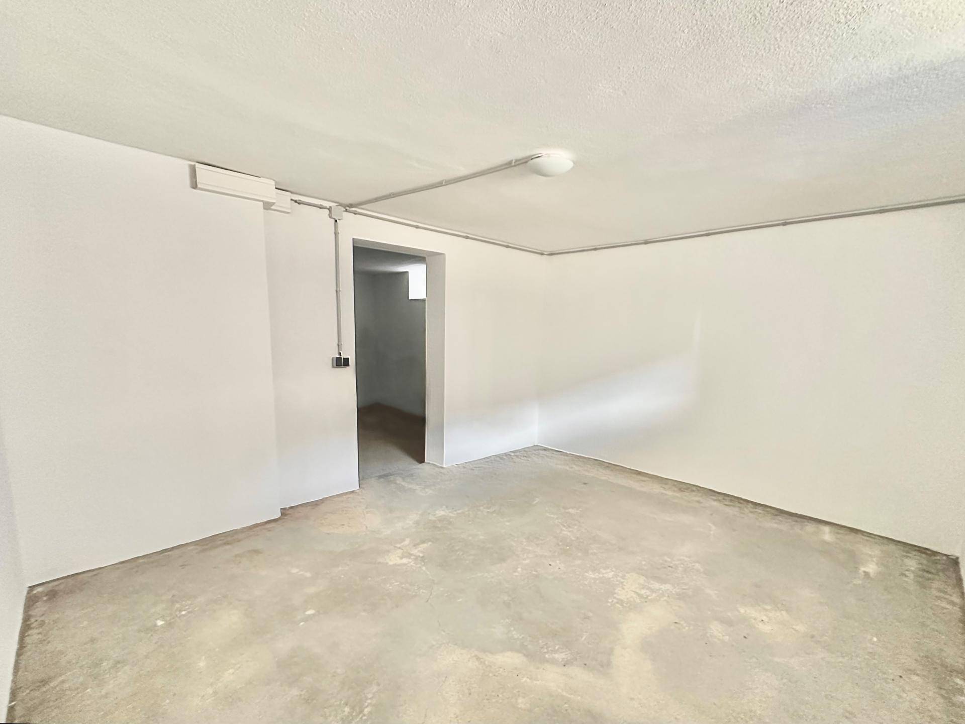Funktionaler Kellerraum mit Betonboden, weißen Wänden und Zugang zu einem weiteren Bereich.