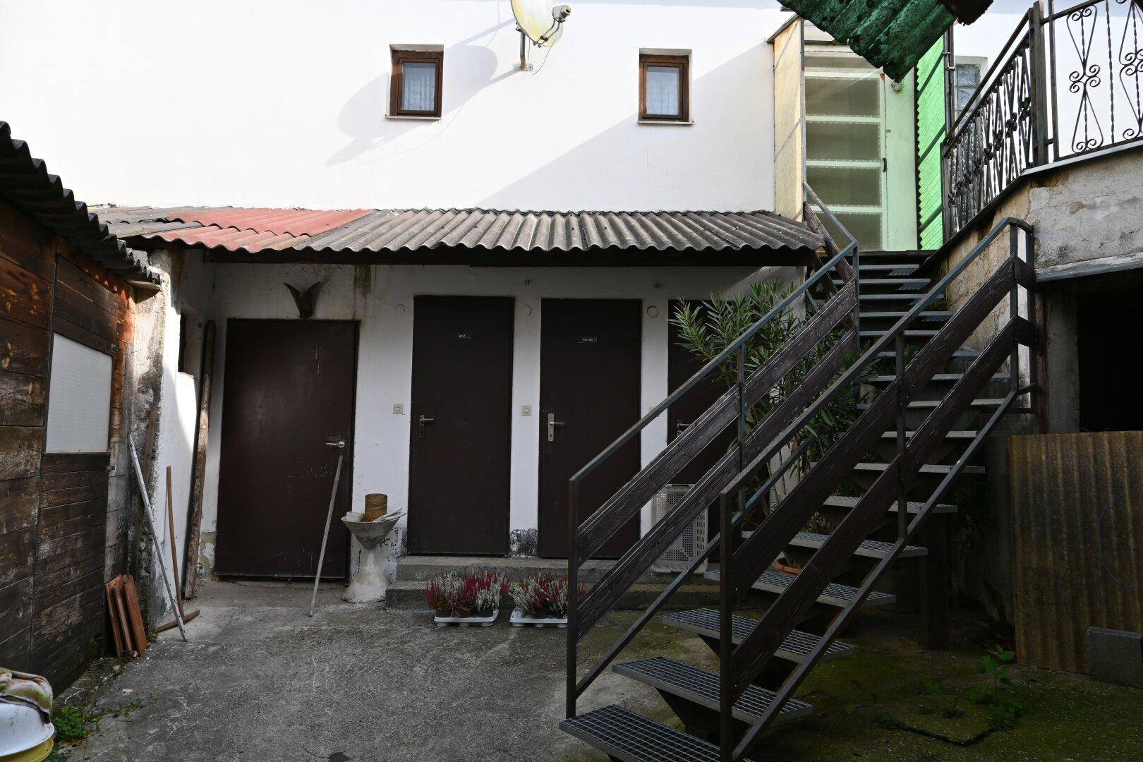 Hinterhof mit Außentreppe, mehreren Türen und einem kleinen Schuppen, der renovierungsbedürftig ist.