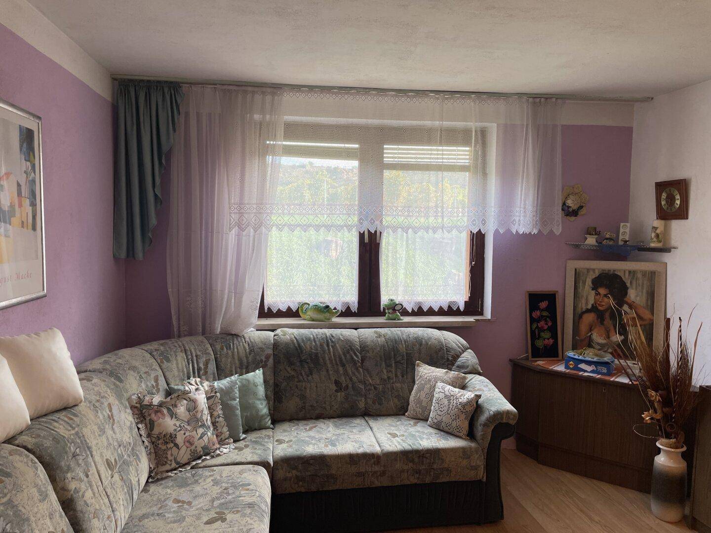 Wohnzimmer mit lila Wänden, Sofa und großem Fenster mit Blick ins Grüne, das renovierungsbedürftig ist.