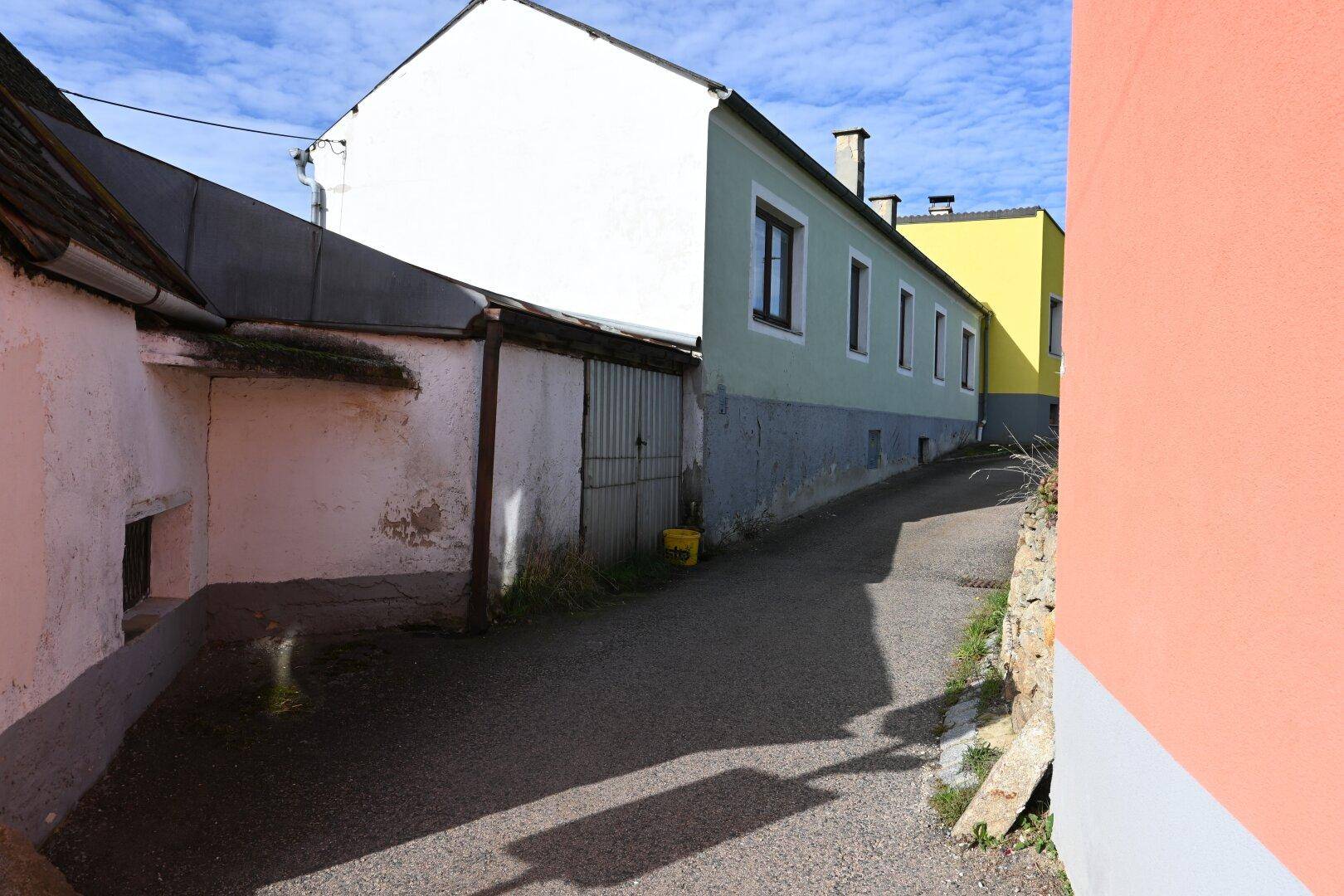 Schmale Gasse zwischen Gebäuden mit unterschiedlichen Fassadenfarben und einem Garagentor, die renovierungsbedürftig ist.