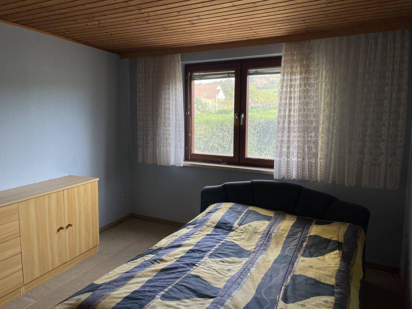 Schlafzimmer mit Holzdecke, blau gestrichenen Wänden und einem Fenster mit Blick ins Grüne, das renovierungsbedürftig ist.