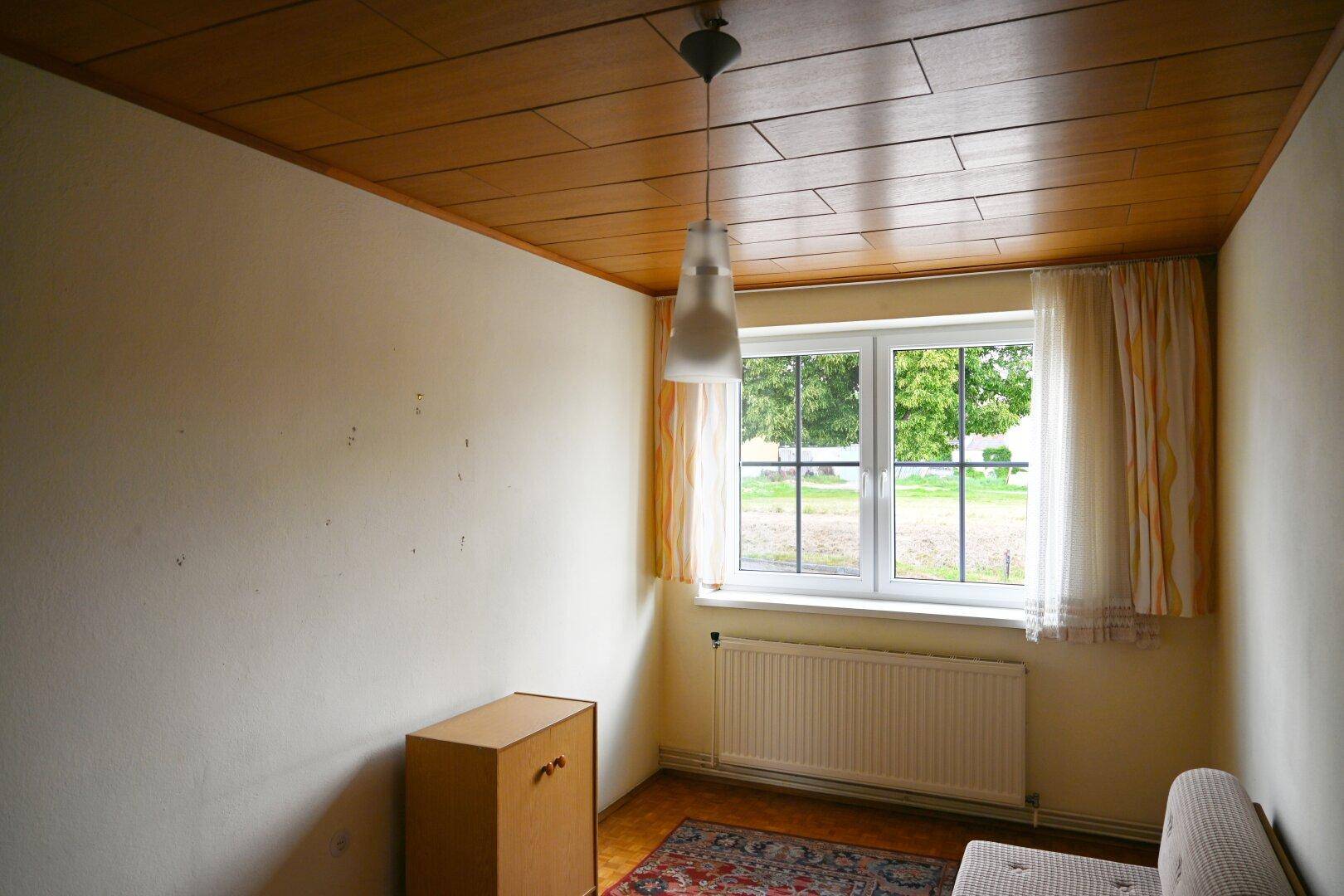 Kleines Schlafzimmer mit Holzdecke, Fensterblick ins Grüne und Heizkörper.