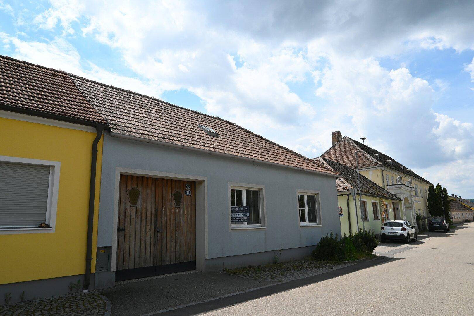 Ländliches Wohnhaus mit hellgrauer Fassade, Holztor und Ziegeldach an der Straße.