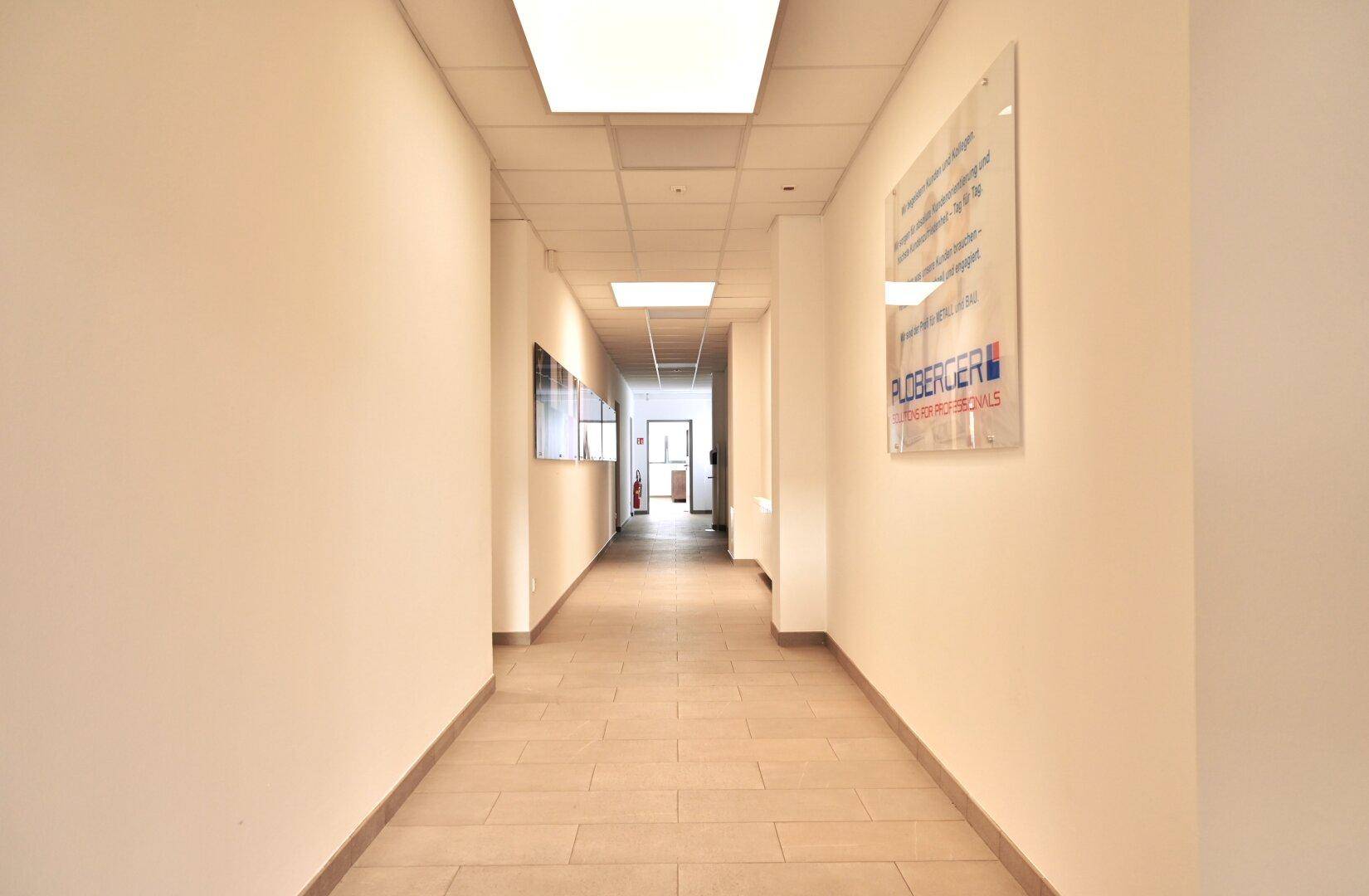 Langer, heller Flur mit Fliesenboden und Deckenbeleuchtung, der zu verschiedenen Büroräumen führt.
