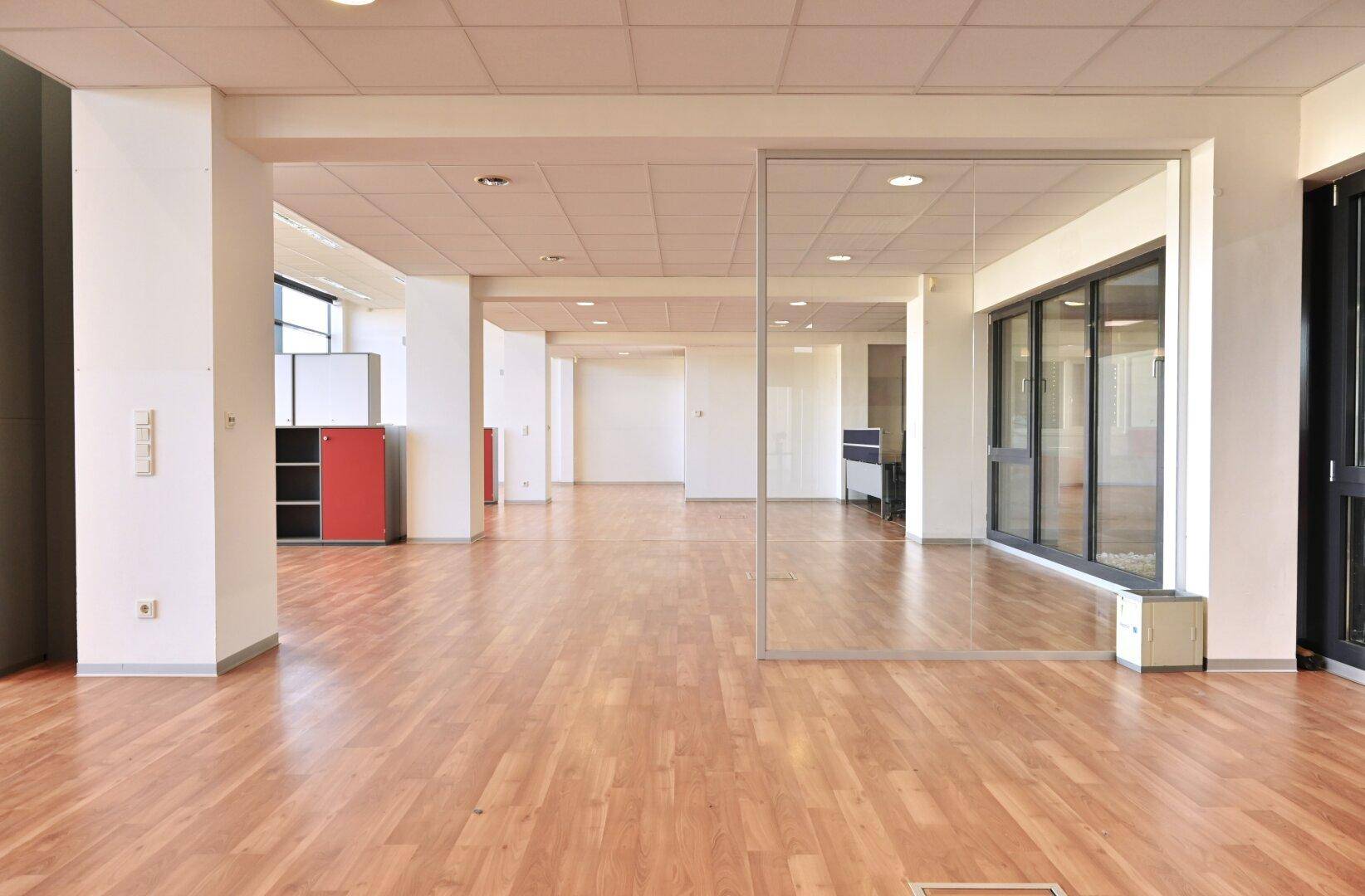 Geräumiger, heller Bürobereich mit Holzfußboden und Glaswand, ideal für flexible Arbeitsplatzgestaltung.