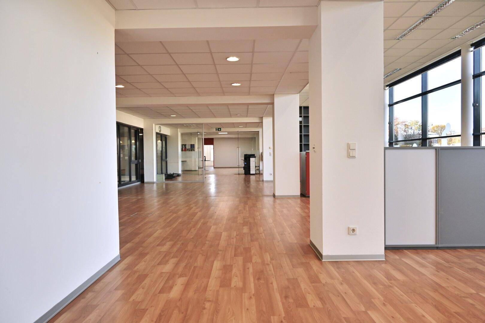 Großer, offener Bürobereich mit Holzfußboden, Säulen und viel Tageslicht durch hohe Fenster.